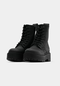 Bottes de combat noires en matériau synthétique texturé, avec un laçage à l'avant, une fermeture éclair sur le côté, et une semelle épaisse à nervures avec des détails de traction.