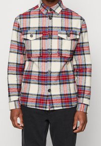 Koszula flanelowa w kratę w kolorach czerwonym, niebieskim i kremowym; posiada dwie kieszenie na piersi, zapięcie na guziki z przodu oraz długie rękawy z guzikami przy mankietach.