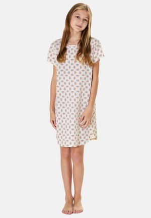 Junges Mädchen steht barfuß und trägt ein weißes Kurzarmkleid mit einem roten und grünen Blumenmuster vor einem einfarbigen Hintergrund.