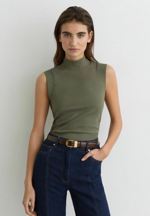 Ung kvinde med lige brunt hår iført ærmeløs grøn rullekrave og mørkeblå jeans med brun bælte, hånd i lommen.