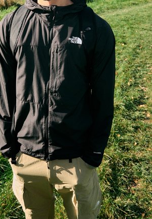 Osoba w czarnej kurtce The North Face i beżowych spodniach typu cargo stojąca na trawiastej powierzchni na zewnątrz.