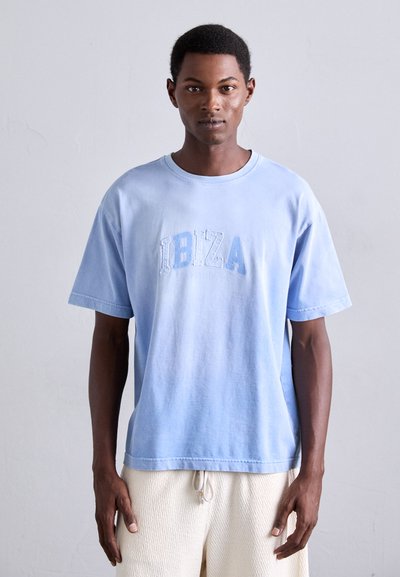 Hellblaues Baumwoll-T-Shirt mit "IBIZA" in dunklerem Blau geprägt, Rundhalsausschnitt, kurze Ärmel und lockere Passform. Getragen mit texturierten cremefarbenen Shorts.