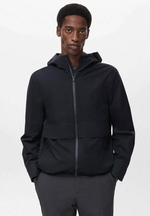 Black hooded jacket with a zip front, featuring a cropped design, soft texture, and side pockets. Worn over gray pants.