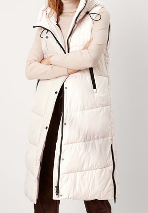 Woman with crossed arms wearing a beige turtleneck and a long white puffer vest with black zippers and snaps, against a plain background.