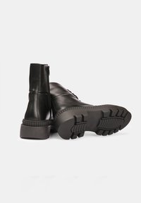 Botas de cuero negro hasta el tobillo con cremallera, suela texturizada y un diseño robusto. Tacón bajo y grueso con un patrón de tracción contrastante en la parte inferior.