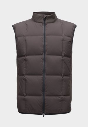 Un homme porte un gilet matelassé foncé par-dessus une chemise blanche à manches longues, avec un pantalon gris et des baskets blanches, sur un fond blanc uni.