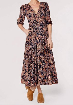 Vrouw draagt een lange, donkere bloemige paisleyjurk met driekwartmouwen en tan suède instapschoenen, staand met één hand in haar zak.