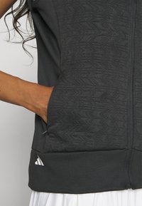 Chaqueta deportiva negra con material texturizado y cierre vertical. Presenta bolsillos laterales y un logotipo blanco de marca en el dobladillo.