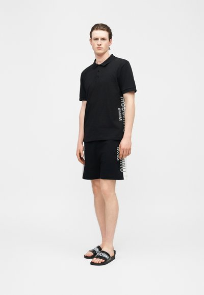 Jeune homme debout portant un polo noir et un short avec un texte sur un panneau blanc, chaussé de sandales noires à enfiler avec un logo blanc.