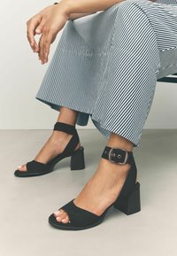 Black block-heeled sandals with ankle straps are worn, showcasing a striped culotte. A hand rests on the leg, emphasizing a relaxed pose.