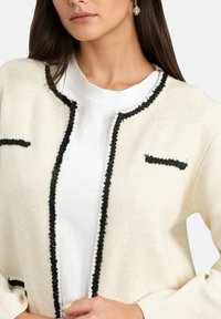 Cardigan crème avec des bordures et des embellissements noirs, doté d'un col rond et de deux petites poches. Porté par-dessus une chemise blanche.