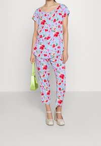 Set in coordinato composto da top e pantaloni in blu chiaro con stampa floreale rossa e rosa, abbinato a scarpe con tacco a blocco beige e una piccola borsa verde.