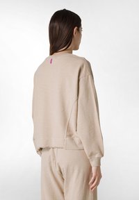 Sudadera de tela suave en color beige con un corte relajado, hombros caídos y una pequeña etiqueta rosa en la parte posterior. Las costuras decorativas añaden textura.
