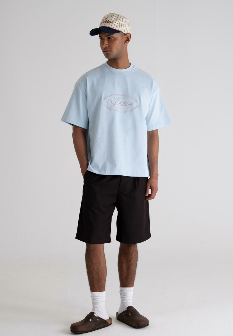 Light blue oversized t-shirt with embroidered logo, black shorts, white socks, tan sandals, and a striped cap with a blue brim.