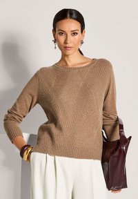Frau in einem braunen strukturierten Pullover, weißen Hosen, mit einer dunkelbraunen Handtasche und mit Ohrringen und Armreifen accessoriert.