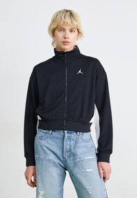 Svart zip-up jacka med hög krage, prydd med en liten vit logotyp p�å bröstet. Bärs tillsammans med ljusblå slitna jeans.