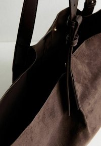 Borsa a mano in suede marrone con manici in pelle scura, apertura superiore e una texture morbida. Presenta un design semplice senza decorazioni visibili.