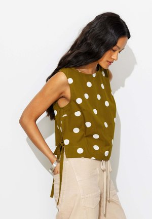 Femme aux longs cheveux foncés porte un haut olive sans manches à pois blancs et un pantalon beige clair à cordon, debout de profil, les mains dans les poches.