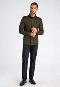 Olijfgroene blouse met lange mouwen en een button-down kraag, zwarte knopen en een getailleerd silhouet. Gecombineerd met donkere, slim-fit broeken.