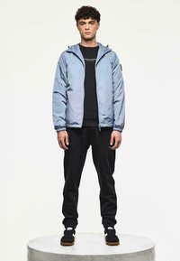 Man die op een rond platform staat, gekleed in een lichtblauwe hoodie, zwarte sweatshirt, zwarte joggingsbroek en zwarte sneakers met witte strepen.