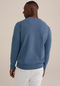 Suéter azul de punto con un acabado texturizado, cuello redondo y puños acanalados, llevado con pantalones de color claro. Características de diseño minimalista.