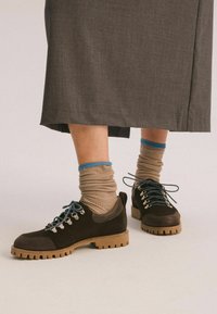 Scarpe in suede marrone con lacci blu e occhielli argentati; suole in gomma con un design testurizzato. Indossate con calze a coste beige con accenti blu.