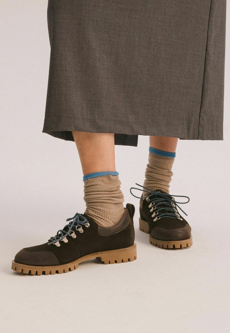 Scarpe in suede marrone con lacci blu e occhielli argentati; suole in gomma con un design testurizzato. Indossate con calze a coste beige con accenti blu.