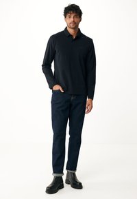 Zwart poloshirt met lange mouwen gecombineerd met donkerblauwe jeans. Model draagt zwarte laarzen met een stevige zool; het shirt heeft een subtiele logo op de borst.