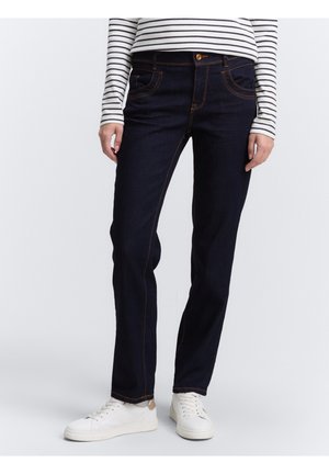 Personne portant un jean droit bleu foncé, des baskets blanches et une chemise blanche à manches longues avec des rayures horizontales noires.