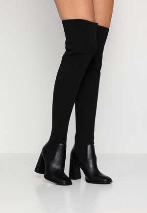 Musta polven yli ulottuvat nilkkurit, joissa on leveä korko, pukeutuneina paljaisiin jalkoihin ja esitettynä yksinkertaisen vaaleanharmaan taustan edessä.