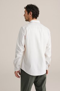 Hvid skjorte med lange ærmer, knapmanchetter, rygstykke og buet kant; kombineret med olivengrønne bukser med struktur. Enkel, skræddersyet design.