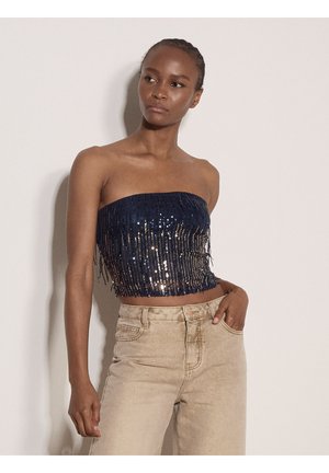 Femme portant un crop top bustier bleu marine à sequins et un jean beige taille haute, debout devant un mur clair uni.
