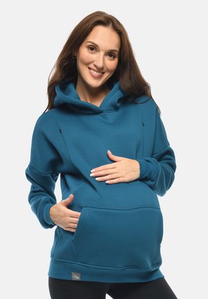 Lächende schwangere Frau in blauem Umstandshoodie mit Hand auf dem Bauch vor einfachem Hintergrund.