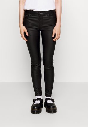 Person trägt schwarze, eng anliegende Hose, weiße Socken, schwarze Schnallenschuhe und ein weißes Hemd, steht vor einem einfarbigen weißen Hintergrund.