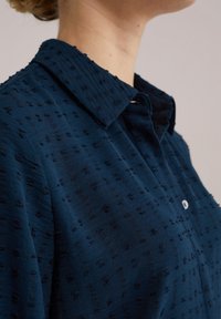 Camisa azul oscuro con un patrón texturizado, cierre de botones y un collar puntiagudo. La tela es suave y tiene detalles en relieve en toda su superficie.