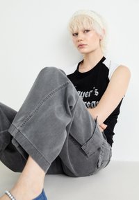 Svart-hvitt grafisk tank topp, grå vid-leggende cargo bukser, og et perlehodebånd. Modell som sitter med armene rundt beina, minimalistisk bakgrunn.