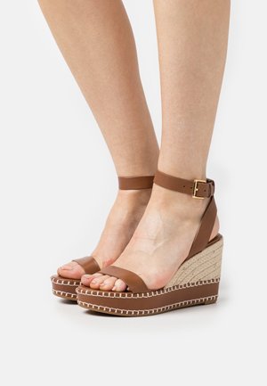 Feet wearing brown leather ankle-strap wedge sandals with rope-textured platform and white stitching on a white background.
