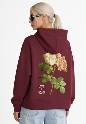 Donna con coda di cavallo biondo platino e occhiali da sole indossa una felpa con cappuccio color bordeaux con rose e la scritta "LOVE & ROSES" sul retro.
