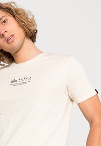Cremefarvet t-shirt med korte ærmer lavet af blødt stof. Har et sort logo og tekst på brystet, samt et lille sort mærke på ærmet.