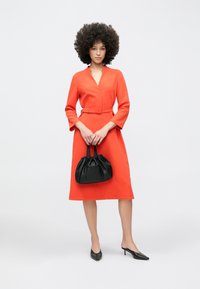 Punane kleit V-kaelusega ja vöökohaga, poolpikkade varrukatega, sobitatud musta voltidega käekoti ja teravate mustade kontsadega. Ühtlane taust.