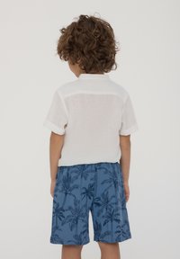 Camicia bianca in lino a maniche corte con polsini arrotolati, abbinata a pantaloni corti blu con motivo a palma. Tessuto liscio, vestibilità casual.