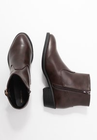 Botas de tobillo de imitación cuero marrón con un diseño curvo, detalle de costura frontal, cierre de cremallera en el costado y un tacón bajo y robusto.