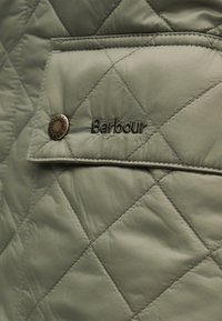 Πράσινο καπιτονέ μπουφάν με λείο ύφασμα, διακοσμημένο με τσέπη και μπρούτζινο κουμπί. Κεντημένο λογότυπο "Barbour" στο ύφασμα.