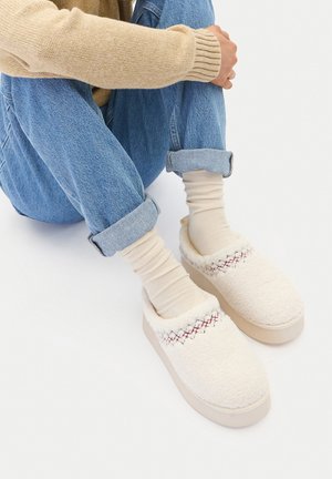 Persona che indossa jeans blu risvoltati, calzini crema e pantofole morbide crema con dettagli di cuciture rosse, seduta con le braccia abbracciate intorno alle ginocchia.