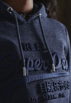 Sweat à capuche bleu marine en tissu texturé avec un logo en relief. Embouts argentés sur les cordons, coupe décontractée et design minimaliste.