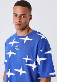 Camiseta de algodón azul con un gran patrón de estrella abstracta blanca, cuello redondo y mangas cortas, con un logo impreso en la parte delantera.