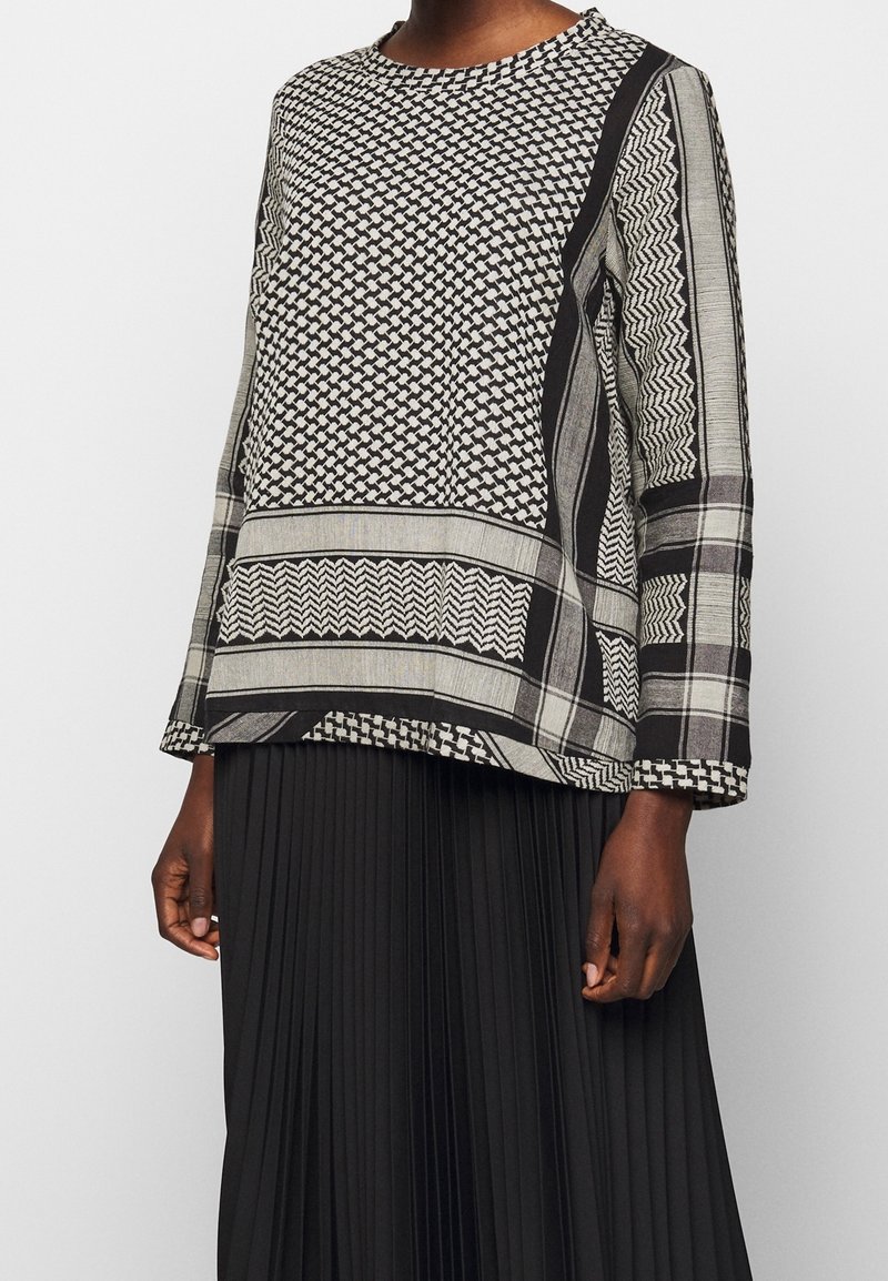 Frau in einer schwarz-weißen, gemusterten Bluse mit langen Ärmeln und geometrischen Designs, kombiniert mit einem schwarzen, plissierten Rock.