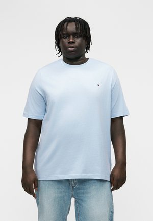 Άνδρας με κότσους ντραντλoκς, ντυμένος με ανοιχτό μπλε T-shirt και ανοιχτό μπλε τζιν, στέκεται μπροστά σε απλό λευκό φόντο.