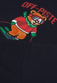 Námořnická mikina s potiskem plyšového medvěda oblečeného v červeném lyžařském obleku, zeleném šále a slunečních brýlích, s nápisem "OFF-PISTE" nahoře.