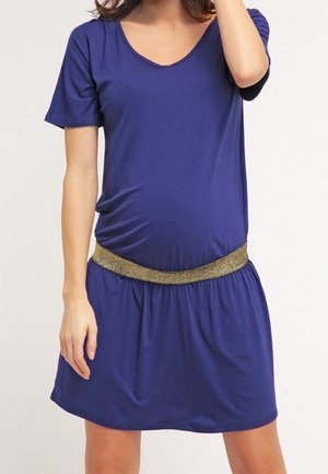 Femme portant une robe bleue à manches courtes, longueur genoux, avec un décolleté arrondi et une ceinture élastique dorée, debout devant un fond uni.